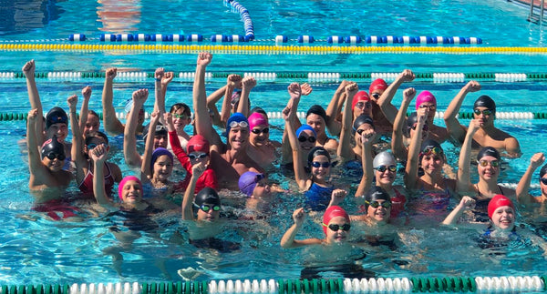 Swimming team poses in the swimming pool raising their hands while wearing Hammer Head Swimming caps.  comfortable, pool, safety, confidence, swim lessons, swim team, coaches, parents, swimmers, head safety, play, pool, underwater, summer 