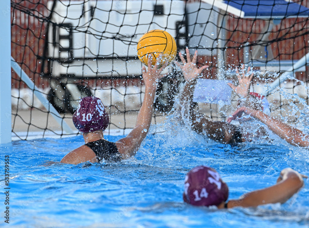 Female water polo players are in action with a yellow ball near a net. This image shows how aggressive players are in the swimming pool. the Hammer Head swimcap is worn under their practice and competition water polo caps for extra head protection to minimize concussions when ball strikes or elbow strikes to the head happen. This will help protect your athletes head.