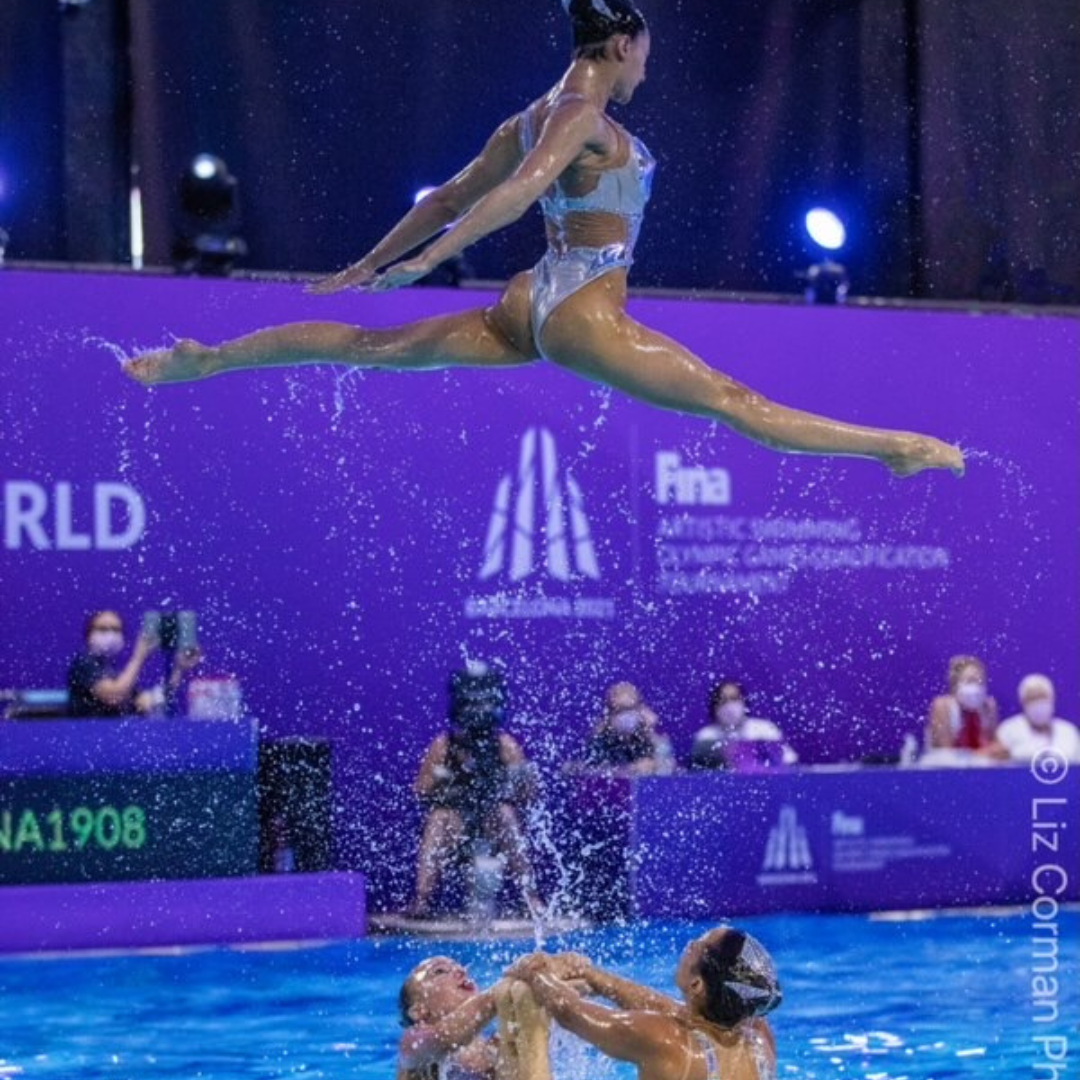 Female artistic swimmer flier (formerly  known as synchronized swimmer) performing a difficult pose feet above the surface of the water. These difficult lifts and throws are practiced repeatedly and frequently end in injury until refined for competition. These athletes often wear a protective swim cap to minimize head injury and keep the team training together as a cohesive team. Hammer Head swim caps are a favorite for concussion prevention.