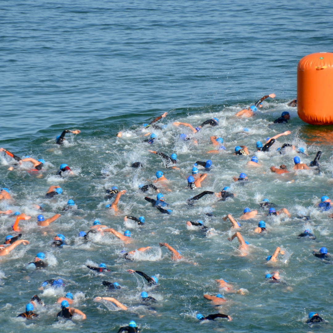 Open water triathlete swimmers in a race with blue swim caps hats. They are approaching the buoy in high traffic with elbows flying as swimmers race to get ahead of each other. This is where head injuries happen all the time. Find the best swim caps for open water swimming. A swim cap with impact protection. Hammer Head is a great solution.